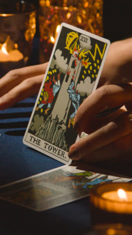 Vertical-Video-Close-Up-Of-Woman-Giving-Tarot-Card-Reading-On-Candlelit-Table-Holding-The-Tower-Card